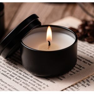 Black matte scented candle in a tin container with a burning flame, placed on an open book