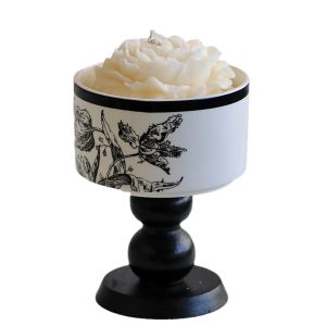 Black pedestal candle holder with white rose-shaped scented candle in a decorative floral-patterned container.