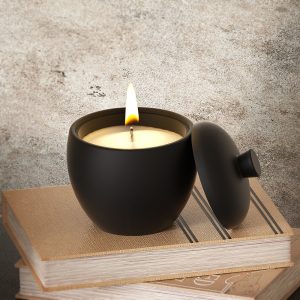 Minimalist matte black scented candle with lid, placed on stacked books, burning with a soft flame.